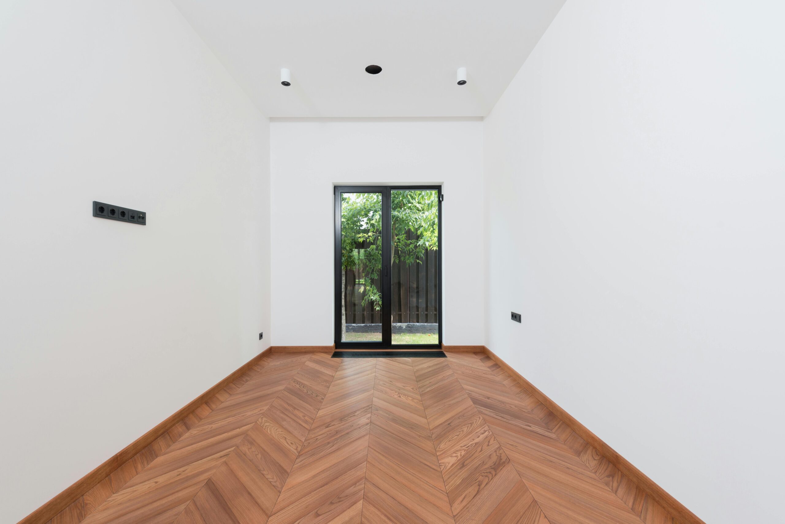 A clean, minimalist interior room with white walls, wooden parquet flooring, and a glass door.